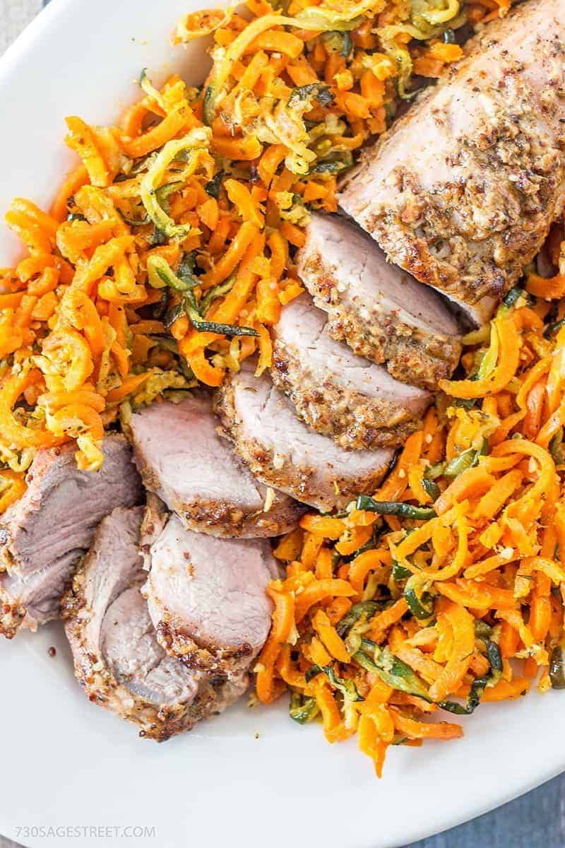 Parmesan Dijon Pork Tenderloin with Garlic herb Veggie Spirals white plate with a sliced pork tenderloin nestled in a bed of spiral carrots and zucchini