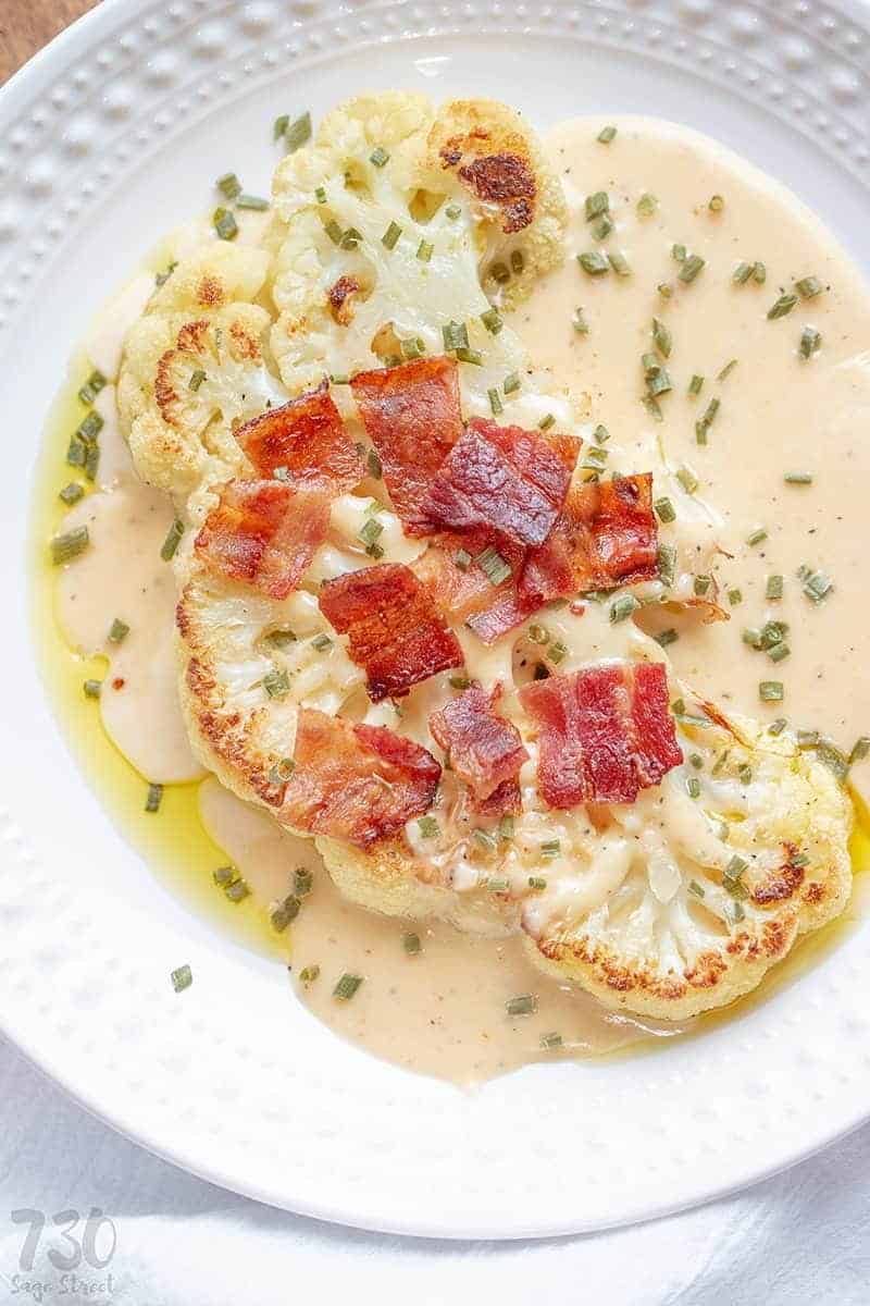 Cauliflower steaks on a white plate with cheese sauce, bacon pieces and chives