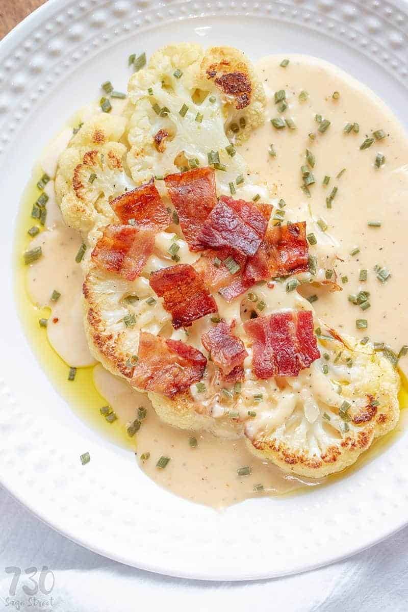 cauliflower steaks on a white plate with cheese sauce, bacon pieces and chives